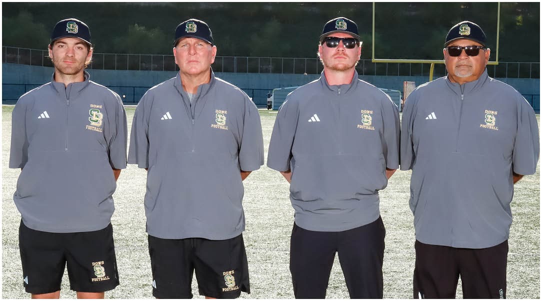 Three adults wearing matching gray sports jackets and caps stand side by side on a sunlit outdoor sports field. Description generated by AI