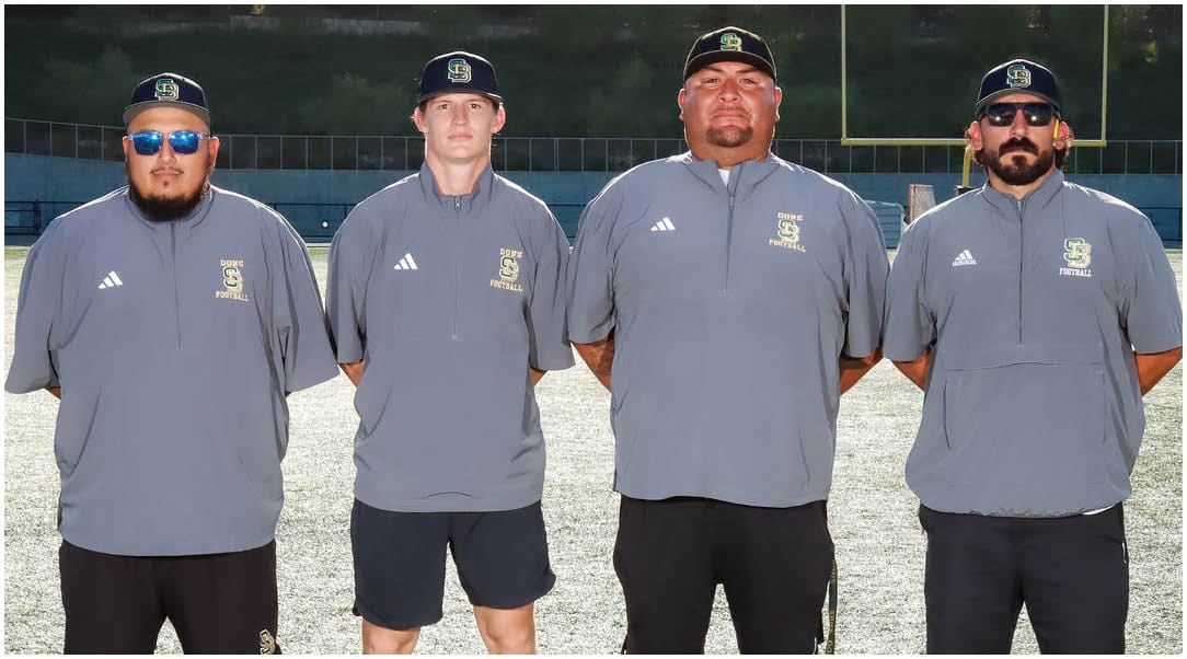 Four adults wearing matching gray sports shirts and caps stand side by side on a sports field with goalposts visible in the background. Description generated by AI