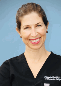 A person with light brown hair tied back is smiling and wearing a black top against a plain blue background. Description generated by AI