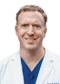 A person with short hair is smiling and wearing a white coat over a blue shirt against a white background. Description generated by AI