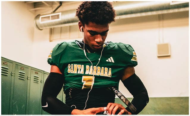 A person wearing a green football jersey with “Santa Barbara" written on it is sitting indoors near lockers, looking down while adjusting their gloves and wearing earphones. Description generated by AI