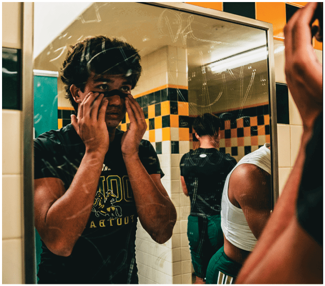 A person wearing a dark sports shirt is standing in front of a mirror in a tiled locker room, touching their face, with two other people partially visible nearby. Description generated by AI