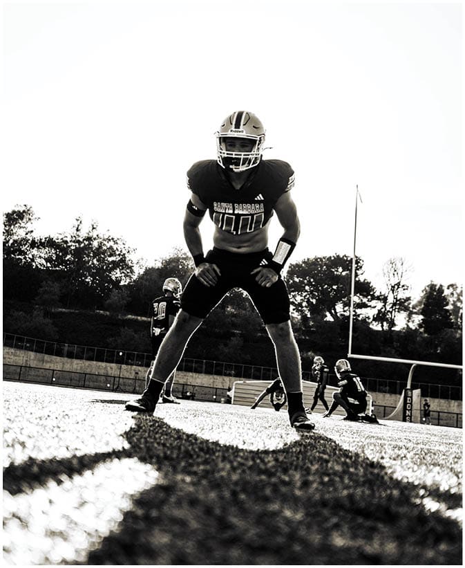 A person in football gear stands in a ready position on a field with other players and a goalpost visible in the background. Description generated by AI