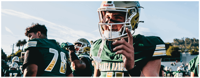 People wearing green and yellow football uniforms are gathered on a field, with one person holding a helmet and another drinking from a bottle. Description generated by AI