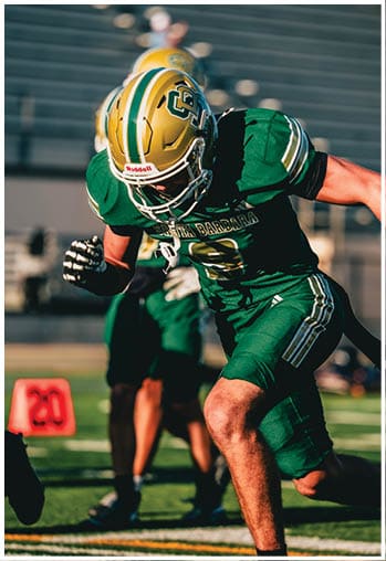An adult in green and gold football gear is running on a field with another player in similar uniform visible in the background near an orange marker. Description generated by AI