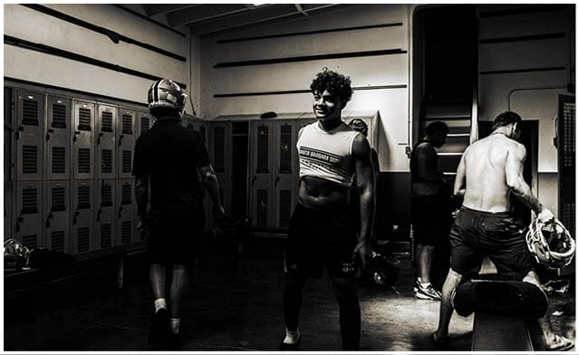 People wearing athletic clothing are walking and standing in a dimly lit locker room with lockers and benches. Description generated by AI