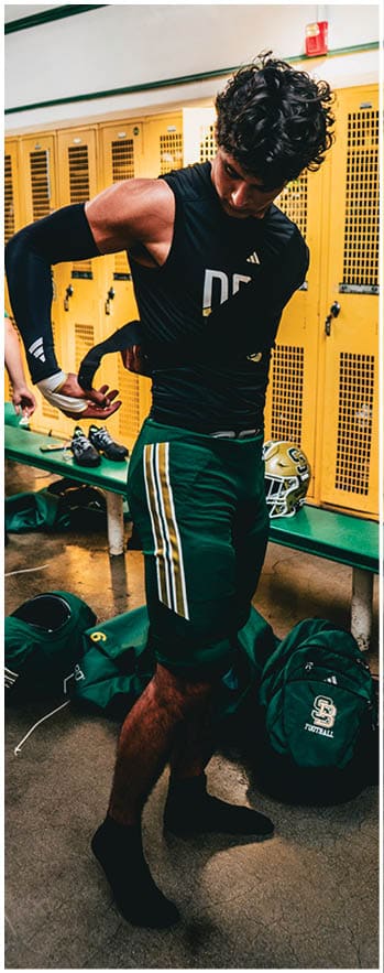 A person wearing a black long-sleeve shirt and green sports pants with yellow and white stripes is standing in a locker room with sports bags and a helmet on a bench nearby. Description generated by AI