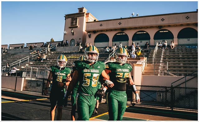 People wearing green and yellow football uniforms are walking on a track near a fenced area in an outdoor sports facility. Description generated by AI