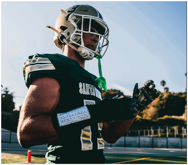 A person wearing a football helmet and green jersey with the number 12 is standing on a field, adjusting their gloves with a play sheet strapped to their forearm. Description generated by AI