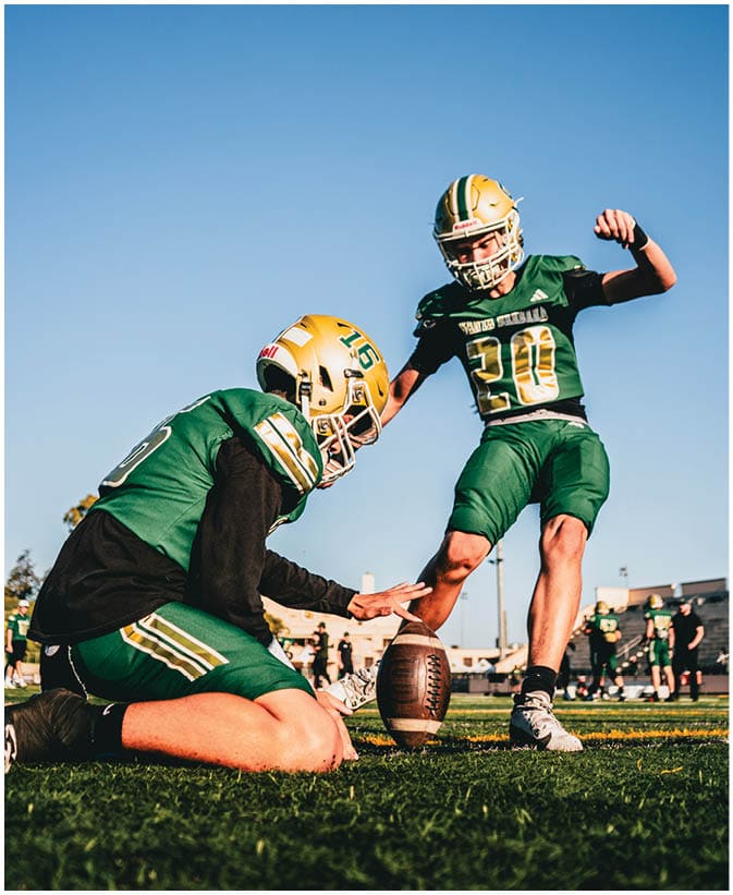 Two people in green and gold football uniforms are on a field with one holding a football upright while the other prepares to kick it. Description generated by AI