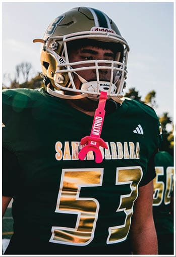 A person wearing a football helmet and green jersey with the number 53 and the word Santa Barbara stands outdoors with a pink mouthguard hanging from the helmet. Description generated by AI