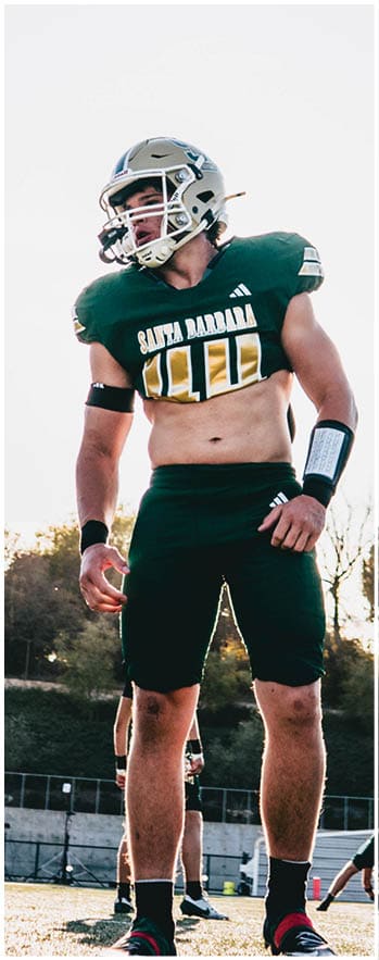 An adult wearing a football helmet and uniform stands on a field with a clear sky and trees in the background while another person is positioned lower behind them. Description generated by AI
