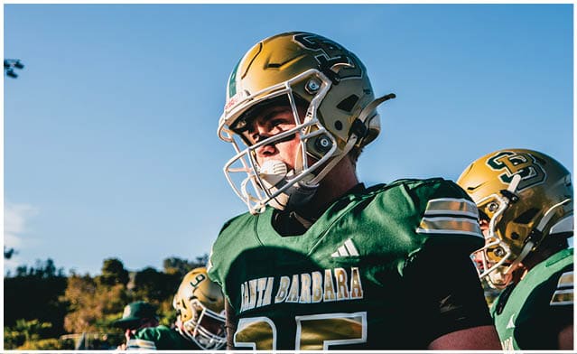 People wearing green and gold football uniforms and helmets stand outdoors under a clear blue sky. Description generated by AI