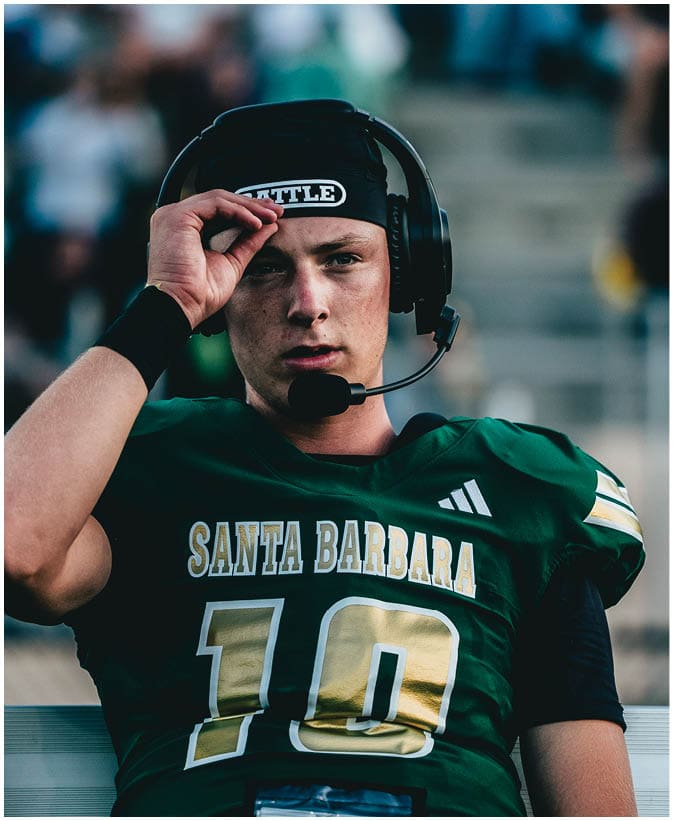 A person wearing a green Santa Barbara football jersey with the number 10 is sitting on a bench and holding a water bottle. Description generated by AI