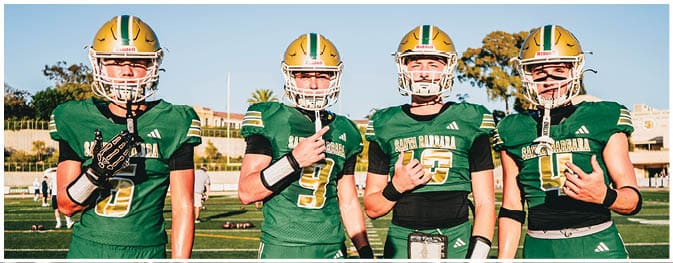 Four people wearing green sports uniforms and gloves stand side by side on a grassy field with visible shadows. Description generated by AI