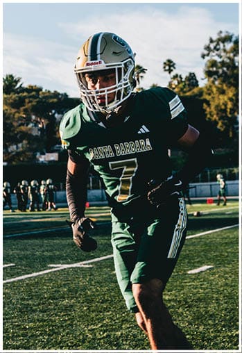 A person wearing a green football uniform with the number seven and a helmet is running on a football field with other players and trees in the background. Description generated by AI