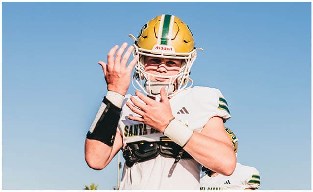 A person wearing a football uniform and helmet is adjusting their gloves on a sunny outdoor field with other players and palm trees in the background. Description generated by AI