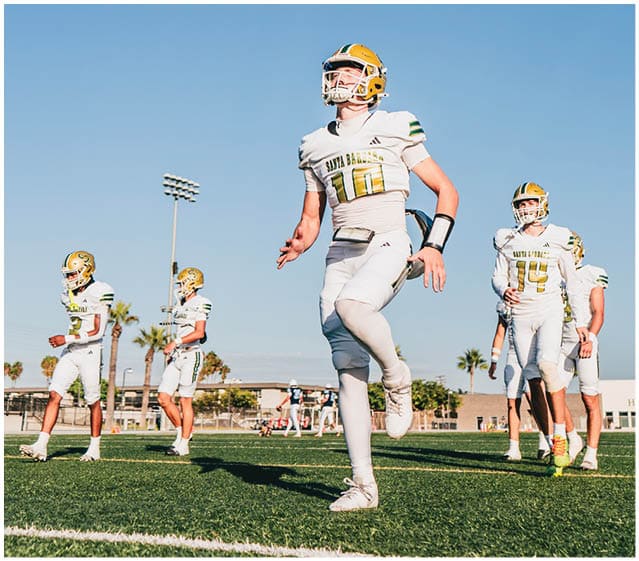 Several people in football uniforms and helmets are standing and walking on a field under a clear blue sky. Description generated by AI