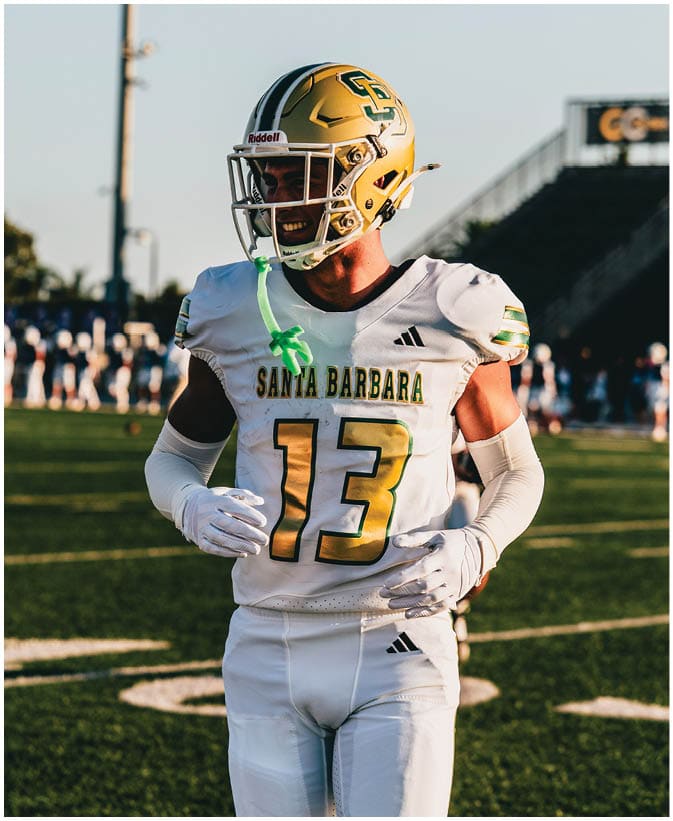 A person wearing a white football uniform with the number 13 and a gold helmet stands on a football field during a game or practice. Description generated by AI