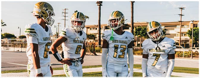 Four people wearing white football uniforms and helmets stand on a grassy field with a parking lot and buildings in the background. Description generated by AI