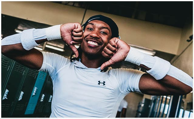 A person wearing a white athletic shirt and wristbands is smiling and posing with both thumbs pointing down in a locker room. Description generated by AI