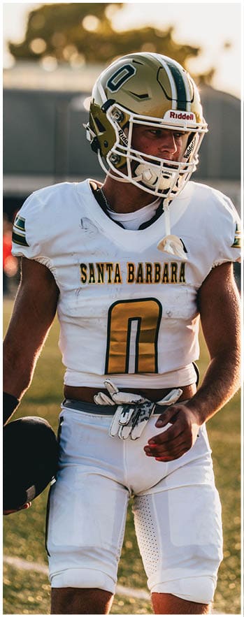 A person wearing a white football uniform with “Santa Barbara" on the jersey and holding a helmet stands on a field during daylight. Description generated by AI