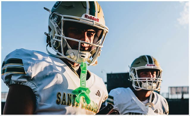 Two people wearing football helmets and white jerseys with “Santa Barbara" written on them are standing outdoors in a stadium. Description generated by AI