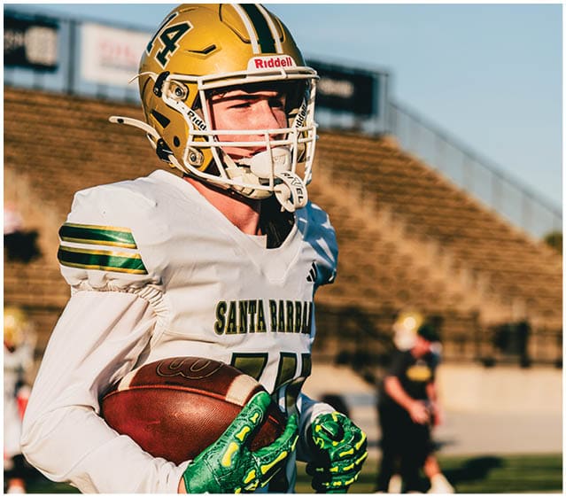A person wearing a gold football helmet and white jersey with “Santa Barbara" on it is holding a football on a field with blurred bleachers in the background. Description generated by AI