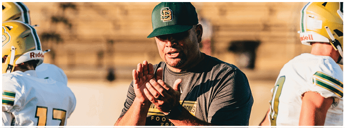 An adult wearing a green cap stands between two people in yellow football helmets on a field with empty bleachers in the background. Description generated by AI