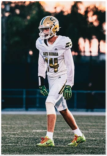 A person wearing a white football uniform with the number 14 and a gold helmet is standing on a field at sunset with trees and poles in the background. Description generated by AI