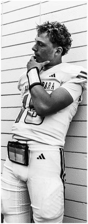 A person wearing a sports uniform with shoulder pads is leaning against a brick wall with one hand touching their chin. Description generated by AI