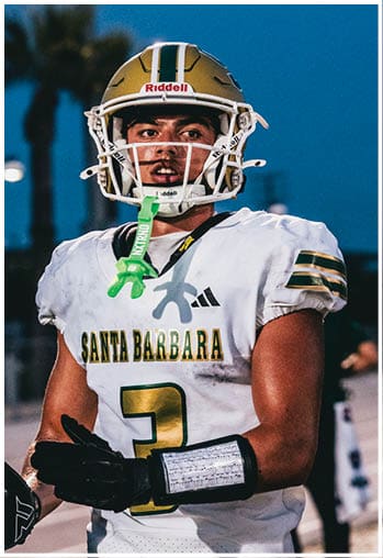 A person wearing a football helmet and a white jersey with “Santa Barbara" and the number three stands on a field at dusk. Description generated by AI