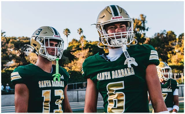 Three adults wearing green and gold football uniforms with helmets stand on a field with trees and a fence in the background. Description generated by AI