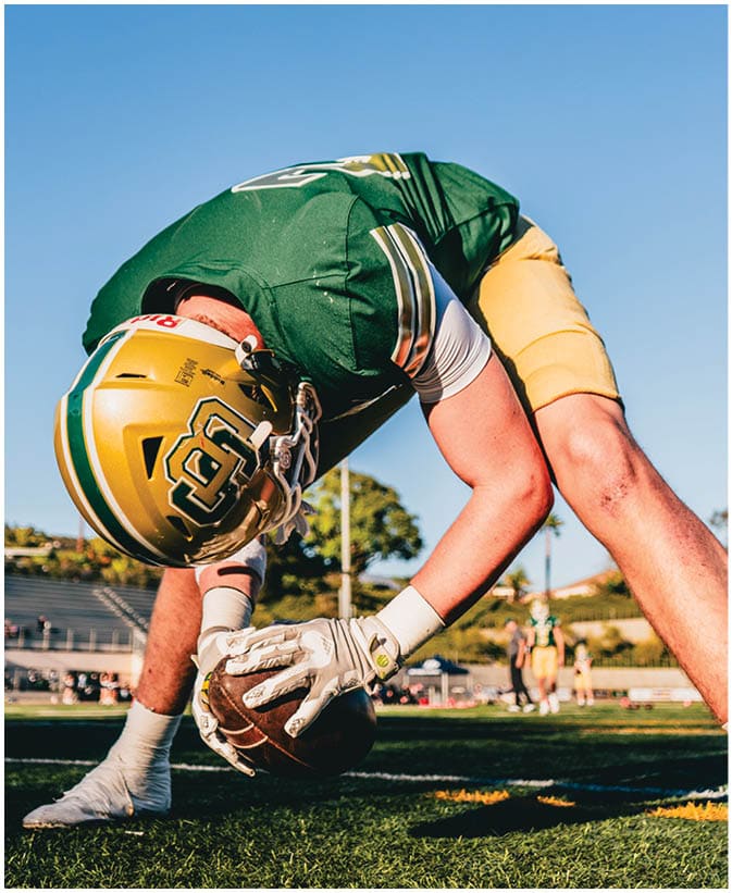 A person in a green and yellow football uniform is bent over on a field, holding a football on the ground as if preparing to snap it. Description generated by AI