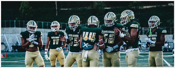 Seven people wearing football helmets and green jerseys are standing together on a field, appearing to be in discussion or preparing for a play. Description generated by AI
