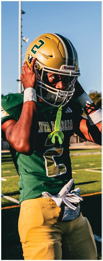 A person wearing a green and yellow football uniform with the number 2 is adjusting their gold helmet on a football field. Description generated by AI