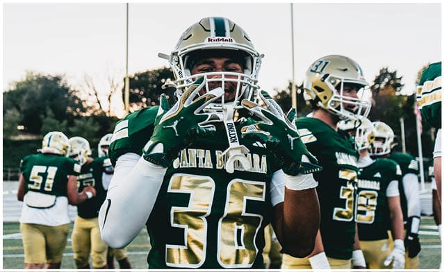 People wearing green and gold football uniforms are standing on a field, with one person in the foreground holding up their gloved hands. Description generated by AI