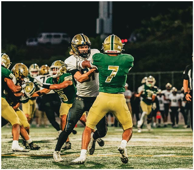 Two teams of people in football uniforms are engaged in a play on a lit field at night with a referee observing nearby. Description generated by AI