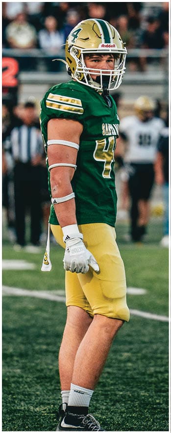 A person wearing a green and yellow football uniform and helmet stands on a football field with blurred people and a scoreboard in the background. Description generated by AI