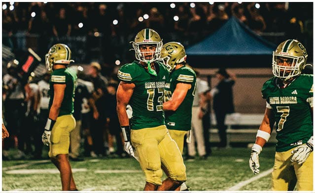 Four football players in green and yellow uniforms stand on a field during a nighttime game with a crowd in the background. Description generated by AI