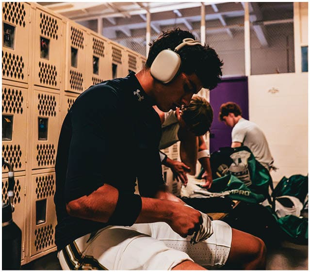 Three people sit in a locker room with one person wearing large white headphones and all appearing focused on preparing or organizing sports gear. Description generated by AI