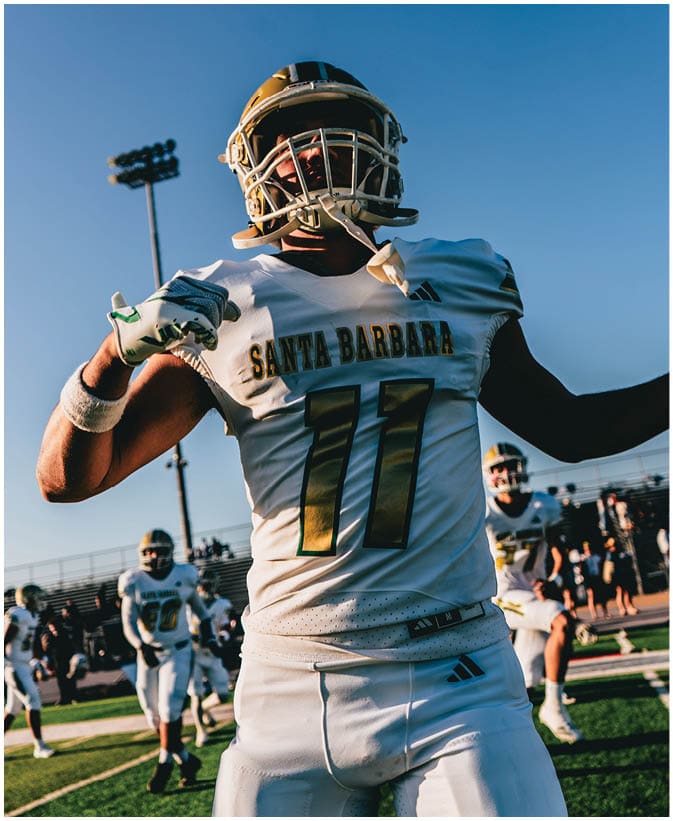 A person wearing a Santa Barbara football uniform with the number 77 is celebrating on a field with other players and a clear sky in the background. Description generated by AI