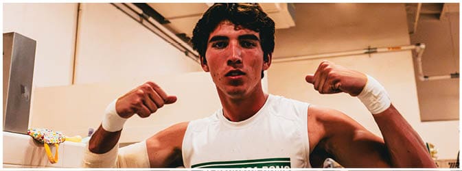A person wearing a sleeveless white shirt and wristband is flexing both arms in a gym or indoor sports facility. Description generated by AI