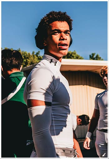 A person wearing a tight white athletic shirt with a black armband and white pants stands outdoors near other people dressed in sports attire. Description generated by AI
