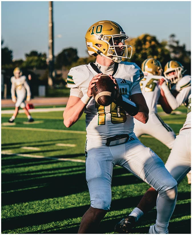 A person wearing a gold helmet and white football uniform is holding a football in a ready-to-throw position on a sunlit football field with other players in the background. Description generated by AI