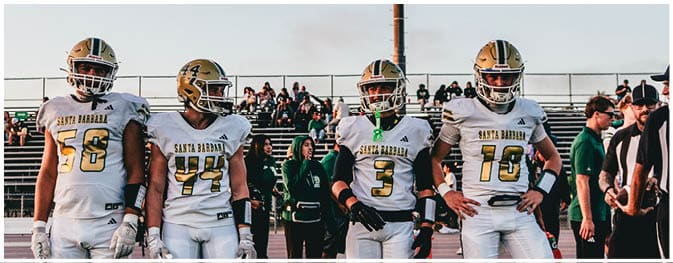 Four people wearing white football uniforms with gold helmets stand side by side on a field with bleachers and spectators in the background. Description generated by AI