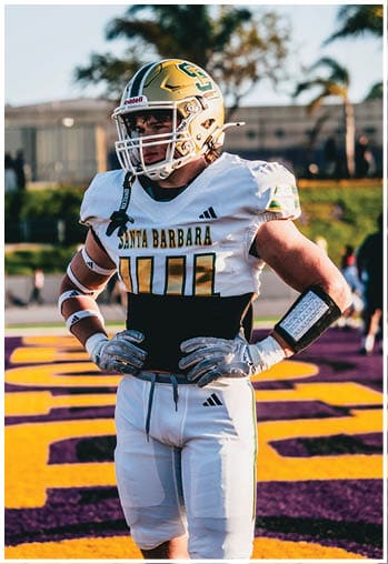 An adult wearing a white and gold football uniform with “Santa Barbara" on the jersey stands on a field with hands on hips, wearing a helmet and gloves. Description generated by AI