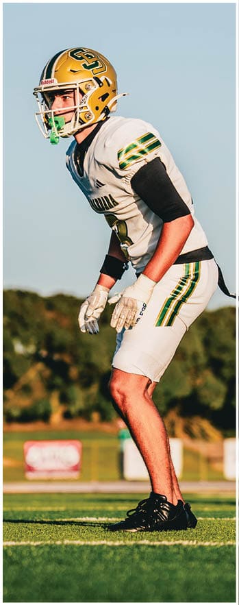 A person wearing a white and green football uniform is standing on a grassy field in a ready position. Description generated by AI