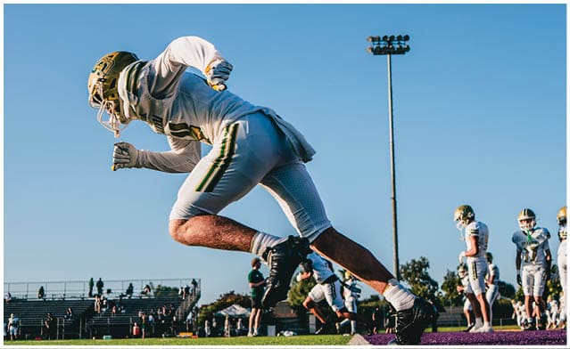 An athlete in football gear is sprinting on a field with other players and spectators visible in the background under a clear blue sky. Description generated by AI