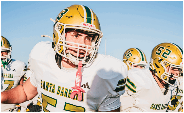 Three people wearing football helmets and white jerseys with green and yellow accents are on a field, with one person in the foreground looking focused. Description generated by AI